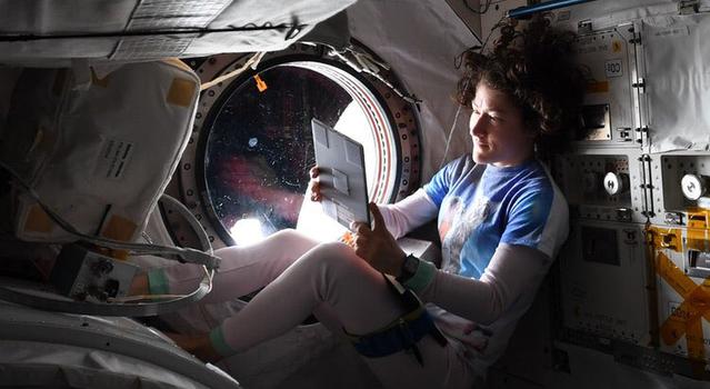 An astronaut looks at her iPad as she relaxes in her bed