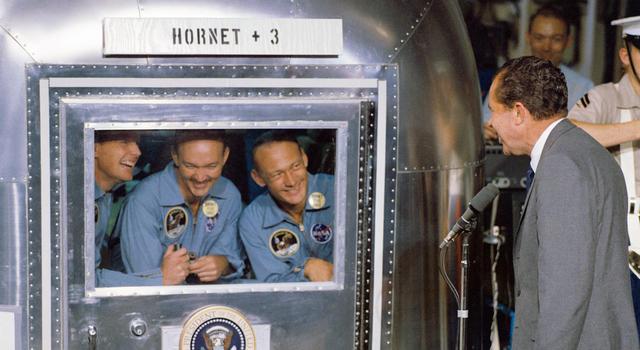 Apollo 11 astronauts stand inside a silver quarantine enclosure while then President Nixon talks to them from the outside.