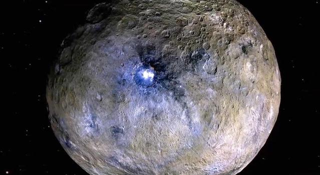 A rocky world covered with craters and bright spots appears brown with splotches of white and purple, the most intense of which is in a large central crater.