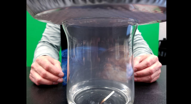 Image of a glass jar partially filled with water and covered with a metal tray. A match is floating in the water and a misty cloud appears to be forming inside the jar.