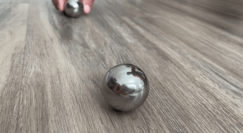 A dense metal sphere is rolled toward another dense metal sphere of the same size and makeup that's sitting still. The sphere at rest rolls forward slowly and the moving sphere continues forward at a much slower pace.