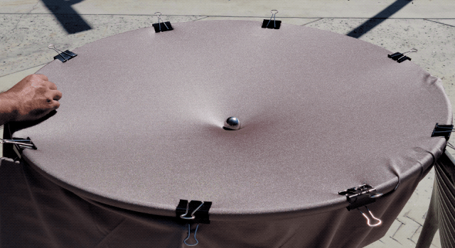 A person releases a marble on a gravity well model made with black material stretched across a hula hoop. A large metal sphere is placed in the center of the model, creating a dip in the fabric.
