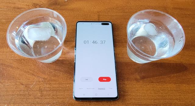 Two clear plastic cups contain one piece of ice melting different amounts. A cell phone timer is set between them.