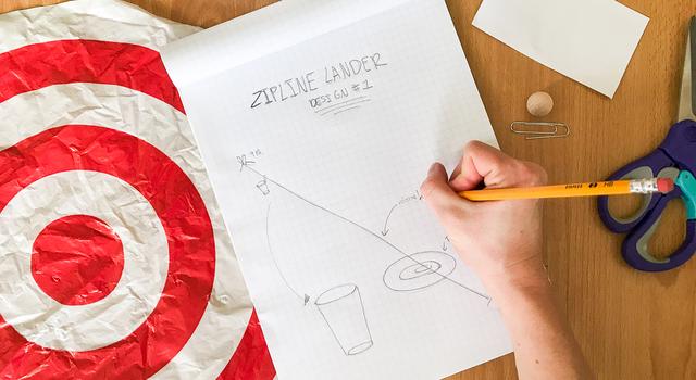 A person sketches a design for their zipline lander on a piece of graph paper with various materials spread around them