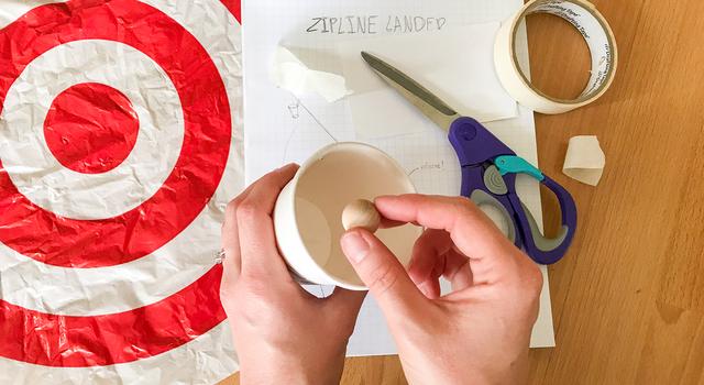 A person puts a wood ball into a paper cup as various materials are scattered about below them