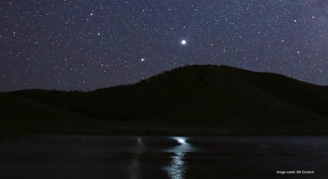 A photograph taken at night showing a lake in the foreground and a hill on the horizon. A starry sky with two bright objects is above the hill.