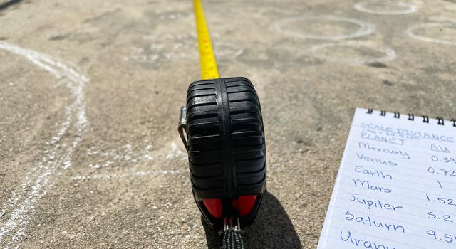 Measuring tape extends on a concrete walkway from an outline of the sun drawn with chalk