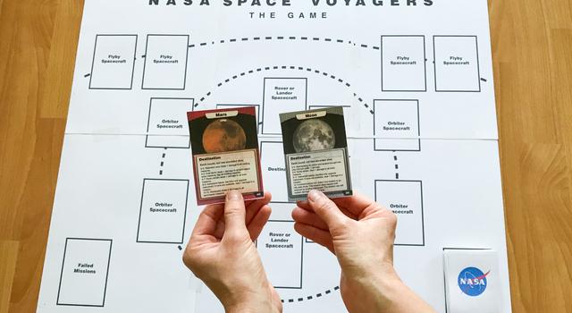 The destination cards for the Moon and Mars are held up over the game mat.
