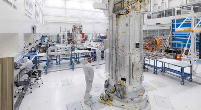An engineer stands in a tall white room to the left of Europa Clipper’s developmental test model, a large, aluminum cylinder with an aluminum box and other aluminum structures protruding from the box.