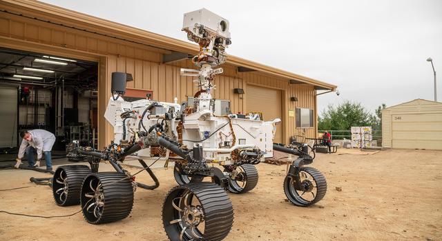 A full-scale working model of the Perseverance Mars rover looks out over a flat sandy area while an engineer in a white coat adjusts a thick tether attached behind the rover.