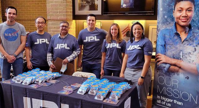 A group of people in JPL shirts poses behind a table with JPL swag.