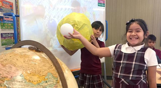 Students hold up models of Earth, the Sun, and the Moon with the Moon between Earth and the Sun.