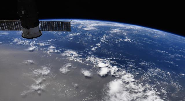 A small piece of the ISS is visible in the top corner of this view looking down from space station over Earth. A large cloud of dust takes half the view over Earth's surface.