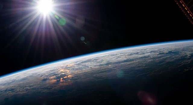 Spikes of light extend from the Sun shining above the glowing blue limb of Earth, as shown from space.