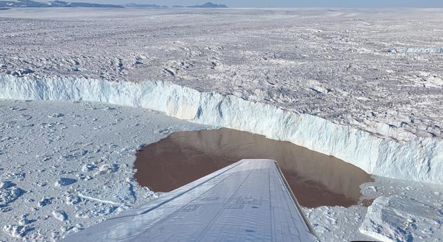 Just beyond the wing of a plane, the edge of a tall glacier is visible through the plane's window. At the bottom of the glacier, bits of ice surround an elliptical pool of brown water at the glacier's edge.