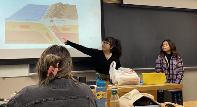 A woman stands at the front of a classroom and points to diagram projected on a screen that depicts tectonic plate activity while a woman stands off to the side and another person is seated facing the front.