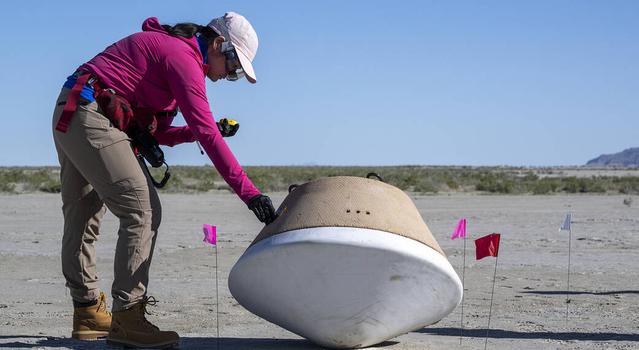 A woman wearing a pink sweatshirt and safety goggles reaches down to touch a large capsule resting on the surface of a desert landscape and surrounded by small flags.