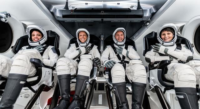 Four astronauts clad in a white suits, hard-shell helmets, and knee-high black boots sit side by side in bucket seats and hold up four fingers.