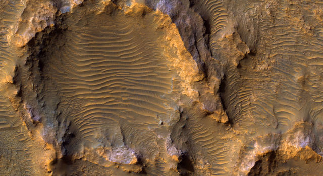 Dunes stretch out across the center of a jagged, rocky crater shown in tan in orange with spots of blue and purple.